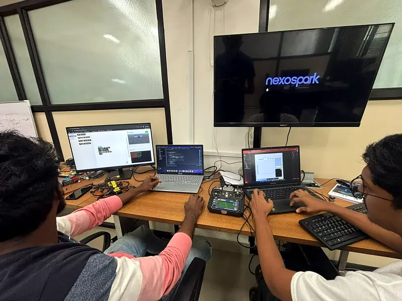 Two Nexospark engineers at the lab desks with a flight simulator, code and a transmitter, the Nexospark logo on screen